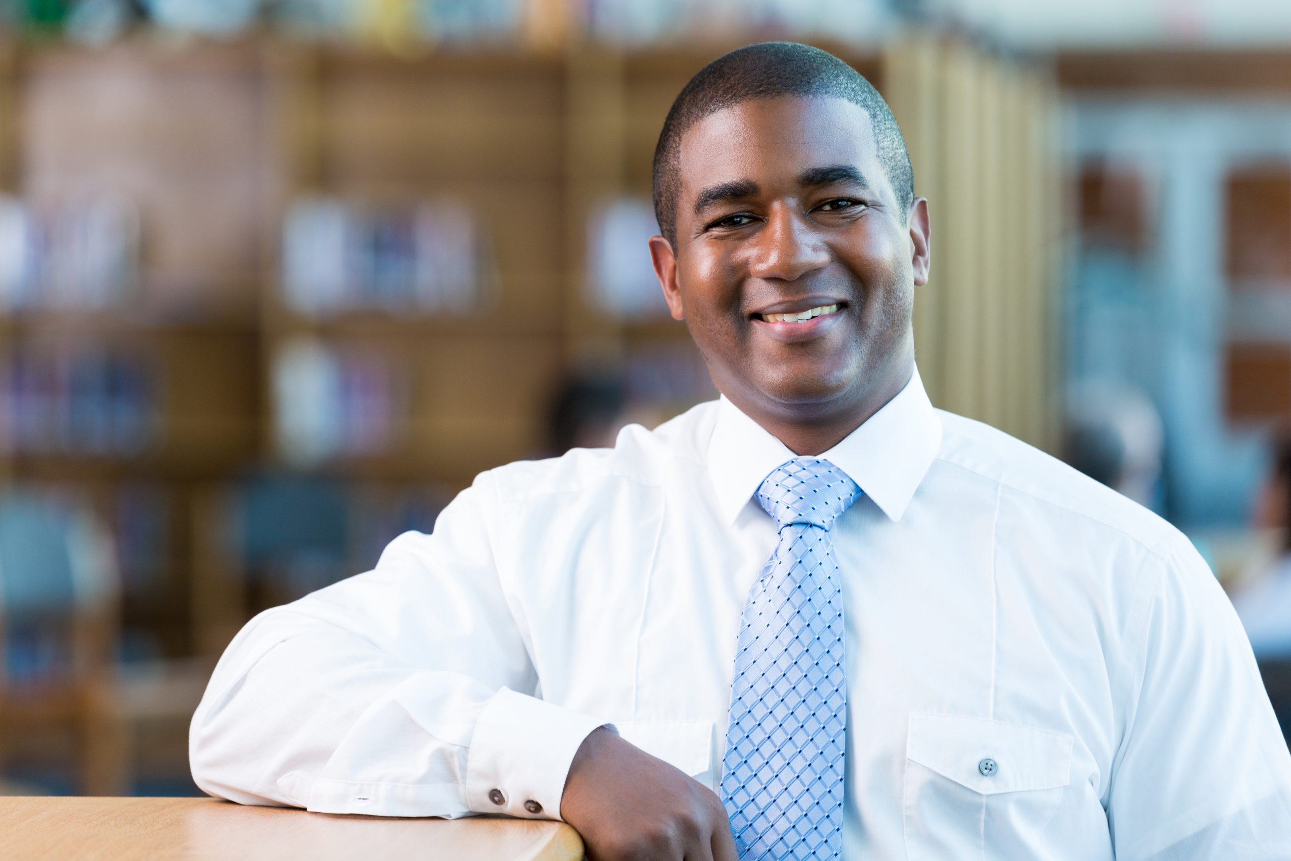 Mid adult African American teacher smiles in high school library. He is leaning on a bookshelf. He is wearing a white button-down shirt a light blue patterned tie.