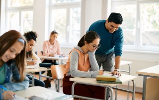 A teacher smiling as he assists a student during class. The student, seated at a desk, looks focused as she works on her assignment, while other students are engaged in their own tasks in the background.