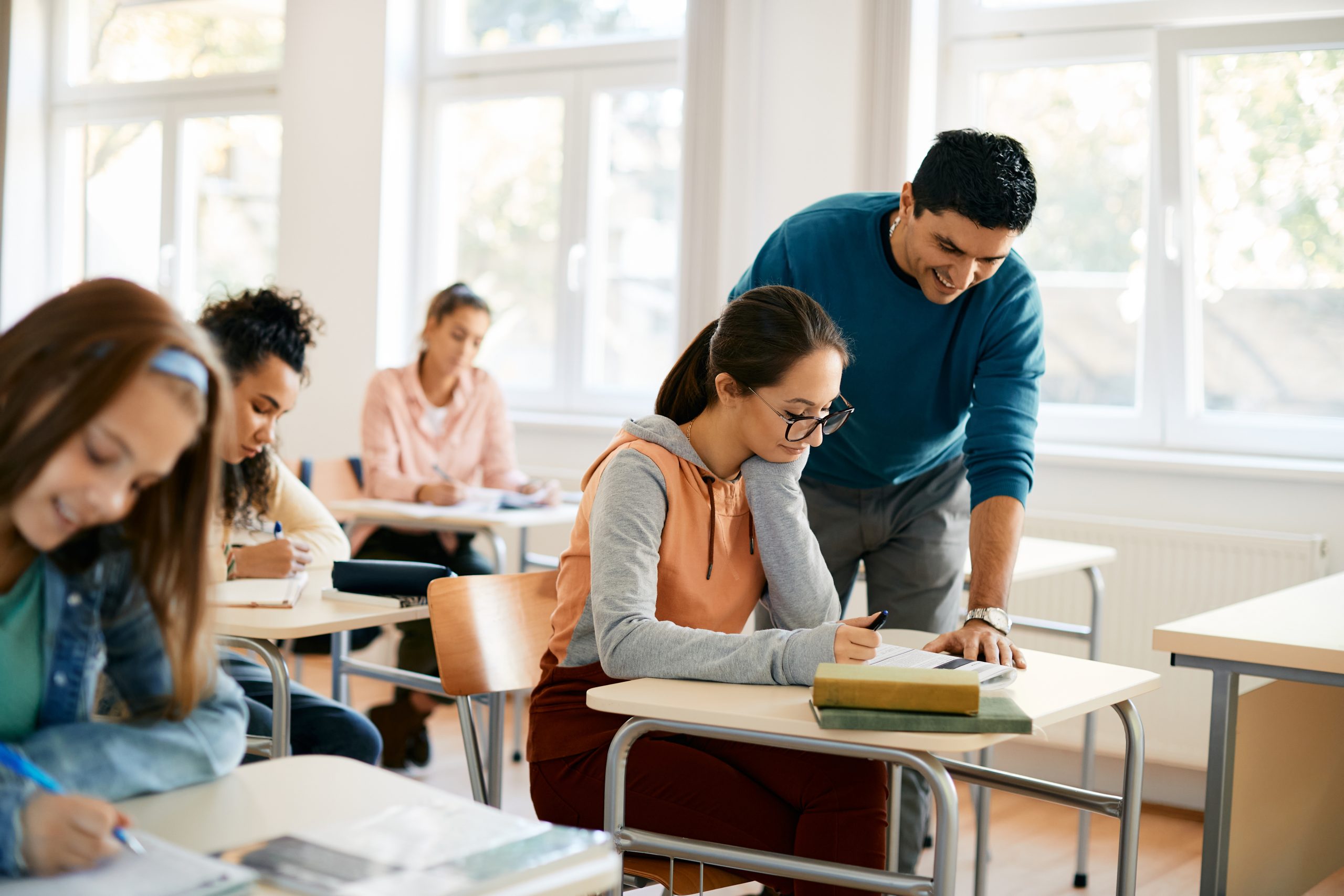 A teacher smiling as he assists a student during class. The student, seated at a desk, looks focused as she works on her assignment, while other students are engaged in their own tasks in the background.