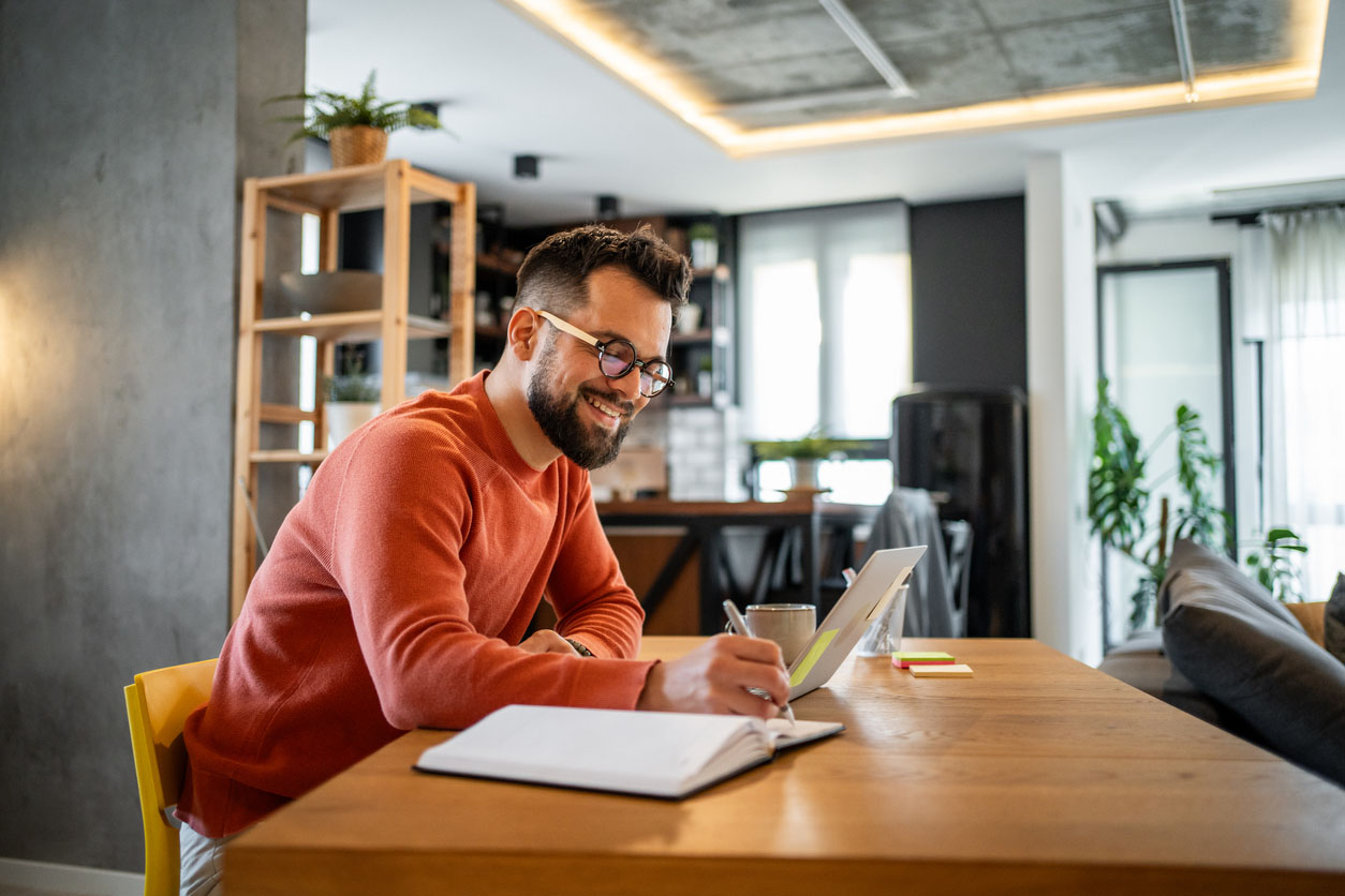 Young man smiling while working from home, seated at a dining table, taking notes in a notebook and reading content on his laptop, creating a productive and cheerful atmosphere